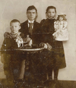 [Translate to Englisch (US):] Schwarz-Weiß-Aufnahme. Grete im dunklen Kleid und mit Puppe im Arm neben ihren Brüdern. Curt im Anzug, Johannes als kleiner Junge im Ringelpullover.